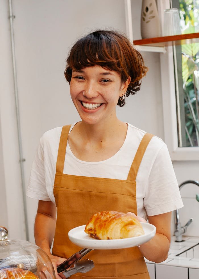 Smiling female baker presenting a delicious croissant in a cozy kitchen setting.